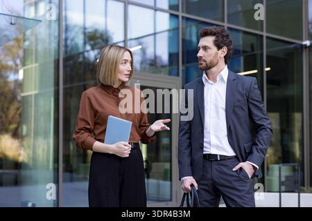 Businesswoman holding a tablet and communicating with a male colleague in a suit, both engaged in an outdoor conversation about work strategies in front of a modern corporate building Stock Photo