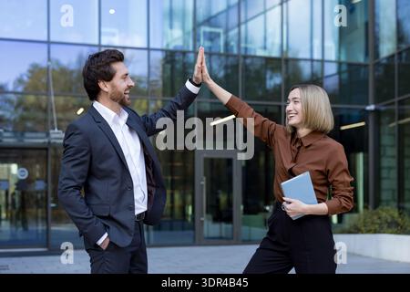 Two smiling multi-ethnic business colleagues standing outdoors in front of a modern office building, high fiving to celebrate teamwork, success, achievement, and partnership Stock Photo