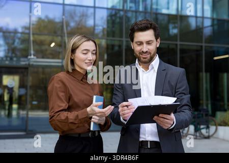 Two business professionals, a woman and a man, are discussing documents on a clipboard while standing outdoors in front of a modern glass office building, collaborating and exchanging ideas Stock Photo