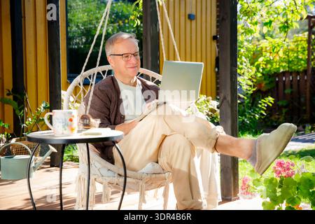 Middle-aged man working remotely from laptop and drinking coffee in hanging chair on sunny patio, relaxed lifestyle and modern home office. Stock Photo