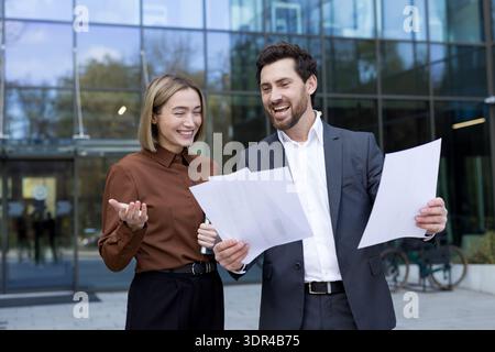 Two smiling business professionals engaging in a lively discussion outside an office building, reviewing paperwork and collaborating on a project with shared enthusiasm Stock Photo