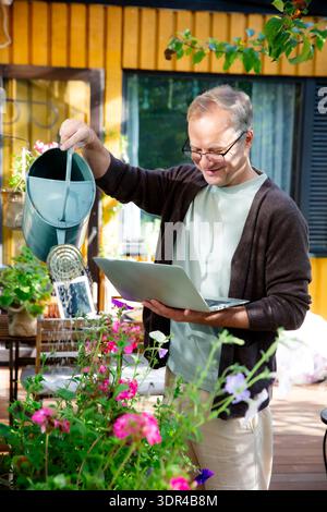 Middle-aged man watering flowers and working remotely on laptop during video call on sunny patio, modern multitasking lifestyle concept. Stock Photo