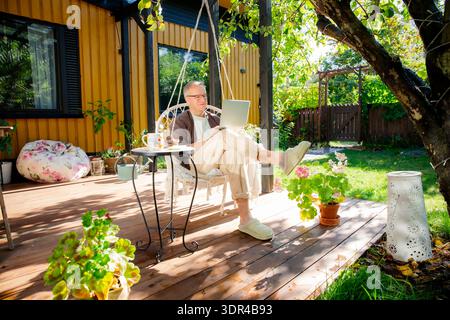 Middle-aged man working remotely from laptop and drinking coffee in hanging chair on sunny patio, relaxed lifestyle and modern home office. Stock Photo