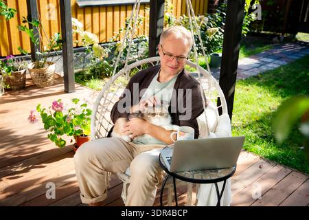 Middle aged man working remotely in swinging chair with laptop and cat on his lap, cozy home office on patio, pet companionship and digital lifestyle Stock Photo