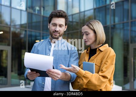 Two business professionals reviewing documents and discussing work, collaborating on a project or an agreement while standing outside a contemporary glass-fronted building Stock Photo