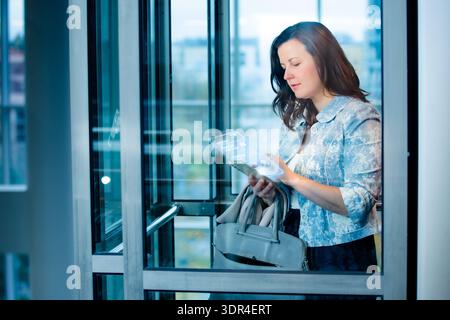 Businesswoman interacting with AI hologram on smartphone inside modern office elevator. Smart building, high-tech  Stock Photo