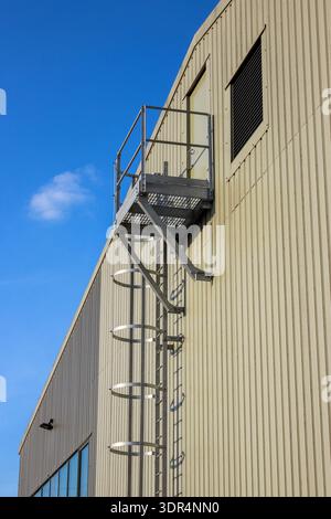 An industrial vertical fire escape ladder with a safety cage and landing platform is mounted on a beige corrugated metal wall. Stock Photo