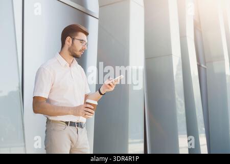 Man Stands by Modern Building While Checking Smartphone and Holding Coffee Cup in Hand Stock Photo