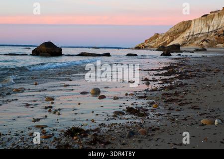 Calm evening seascape with gentle waves, scattered pebbles and seaweed on sandy beach beneath soft pink and blue sky. Stock Photo