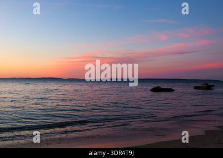 Soft pink and blue sunset sky reflecting over calm sea with gentle waves and distant shoreline silhouettes. Stock Photo