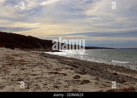 Sandy and rocky beach shoreline with gentle waves, autumn forested cliffs and dramatic cloudy sky over sparkling water. Stock Photo