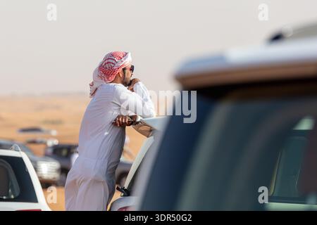 Dubai, United Arab Emirates-March 9, 2025: Local off-road tradition as Emirati man wearing kandura and keffiyeh leaning on SUV in desert, cultural mom Stock Photo