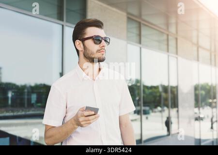 Man Stands Outside Modern Building Holding Smartphone and Wearing Sunglasses in Sunny Weather Stock Photo