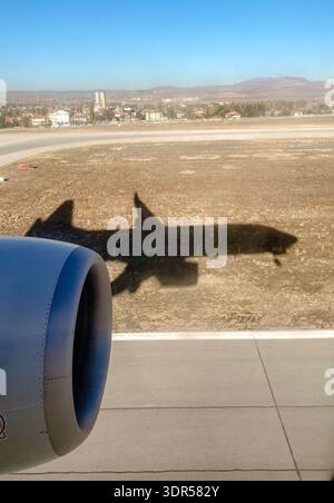 Airplane shadow on runway during landing, view from passenger window with jet engine and dry landscape in background. Stock Photo