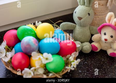 A straw basket filled with colorful Easter eggs sits next to plush rabbit toys. holiday decoration, spring symbols, festive celebration. Easter. Stock Photo