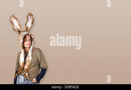 Sstudio portrait of a middle aged woman wearing handmade felted bunny hat with long ears standing against taupe background. Copy space Stock Photo