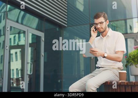 Man Using Tablet and Talking on Phone While Sitting Outside Modern Building in City Stock Photo