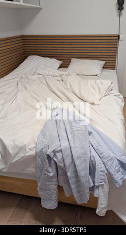 Vertical unmade bed with white bedding in cloud dancer color of the year 2026, blue striped shirt hanging on wooden bed, cozy real bedroom interior in Stock Photo