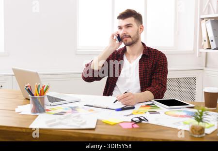 Young Man Working at a Desk With a Phone While Managing Various Tasks in a Bright Room Stock Photo