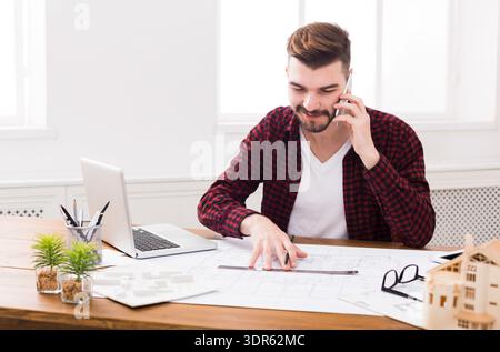 Man Talking on Phone While Working on Architectural Plans at His Desk in Daylight Hours Stock Photo