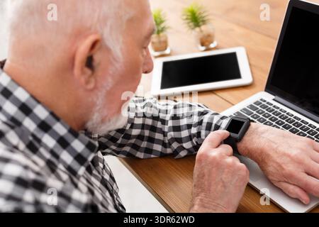 Senior Man Checking Smartwatch While Working on Laptop at Home Office Stock Photo