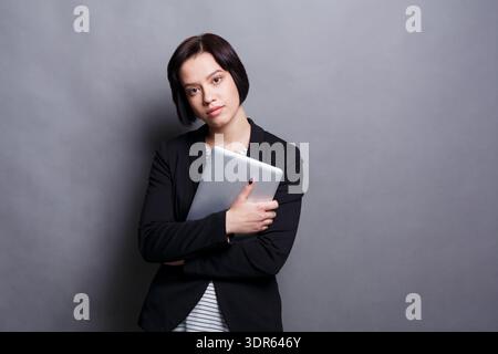 Young Woman Holds Tablet While Standing Against Grey Background in a Casual Manner Stock Photo