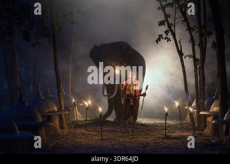 Mahout standing in front of an elephant in an illuminated forest, Chang village, Surin province, Thailand Stock Photo