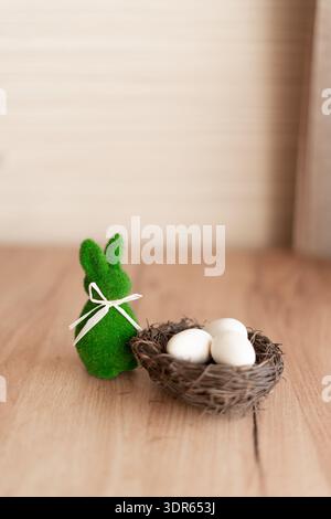 Easter bunny figurine with nest and eggs on wooden background. Green fuzzy Easter bunny figurine with white ribbon beside a small nest holding three Stock Photo