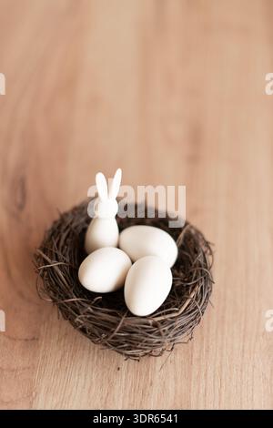 White Easter Eggs and Bunny in Twig Nest on Wooden Background. Minimalist Easter still life: small white bunny figurine and three white eggs in a twig Stock Photo