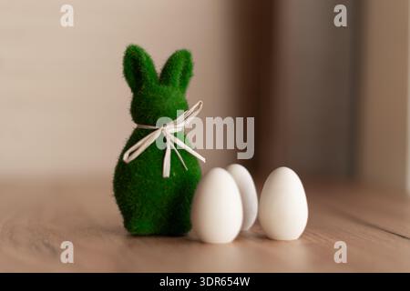 Minimalist easter bunny with eggs on wooden table. Green fuzzy bunny figurine with ribbon beside three white eggs on wooden table, soft focus Stock Photo