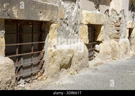 The weathered facade of an ancient structure features rough-hewn stone, crumbling plaster, and rusty metal bars protecting small, dark openings Stock Photo