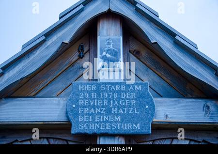 Wooden memorial plaque on alpine gable with black-and-white portrait above; blue-toned weathered wood; engraved german text about a hunter who died in Stock Photo