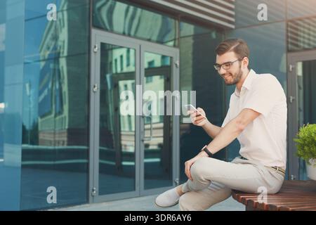 Young Man Checks His Phone While Sitting Outside a Modern Building on a Sunny Day Stock Photo