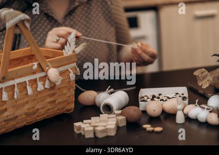 Woman crafting handmade Easter decorations with eggs and basket. Woman crafting Easter decorations at table with wicker basket, eggs, twine, wooden Stock Photo