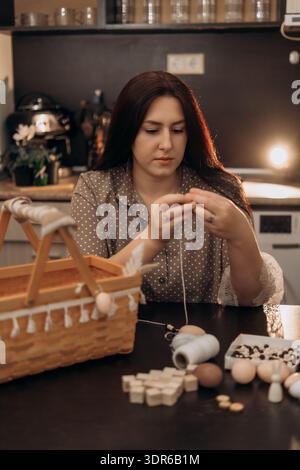 Woman crafting with thread and supplies at cozy home table. Woman doing crafts at kitchen table with wooden basket, thread and small items in warm Stock Photo