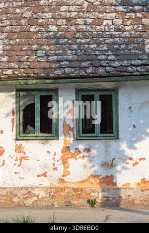 Abandoned old house facade with peeling paint, broken windows and a weathered tiled roof conveying decay, neglect and rustic charm in soft daylight. T Stock Photo