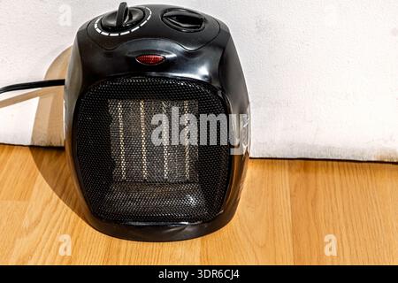 A small black portable electric space heater placed on a wooden floor against a light wall. Home heating concepts Stock Photo