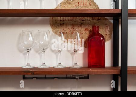 Minimal interior detail showing four empty wine glasses lined up on a rustic wooden shelf beside a translucent red glass bottle, set against a white w Stock Photo