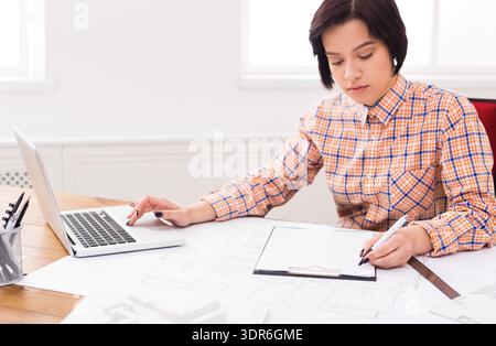 Young Woman Works on Design Plans at Desk With Laptop During Daytime in Office Environment Stock Photo
