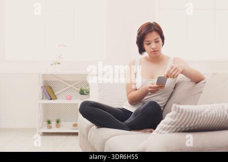 Young Woman Sits on Couch and Checks Phone While Relaxing at Home Stock Photo