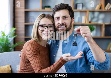 Smiling young adult couple sitting on a sofa and holding up new house keys, celebrating moving into their first home or apartment, representing real e Stock Photo