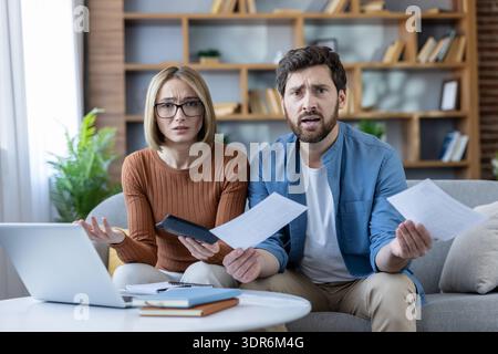 Young couple sitting on a sofa, looking shocked and frustrated while calculating expenses and holding bills, facing financial difficulties, budget iss Stock Photo