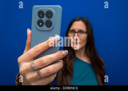 Woman holding a modern smartphone with multiple cameras, taking a selfie against a vibrant blue backdrop Stock Photo