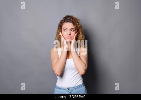 Young Woman Standing Against Gray Wall With Hands on Face Looking Thoughtful Stock Photo