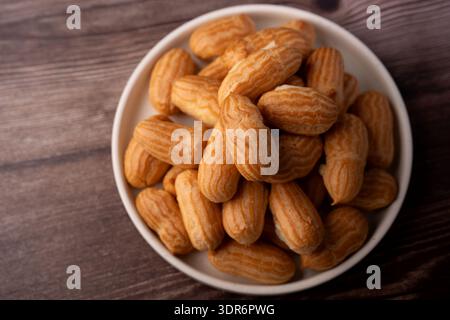 overhead close-up of a rustic white bowl piled with golden-brown mini eclairs on a warm woodgrain tabletop, soft lighting highlighting ridged choux te Stock Photo