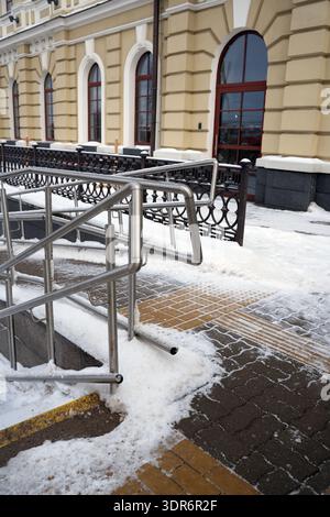 snow-covered stainless-steel handrails and accessibility ramp in front of a beige historic building with arched red-framed windows, ornate black iron Stock Photo