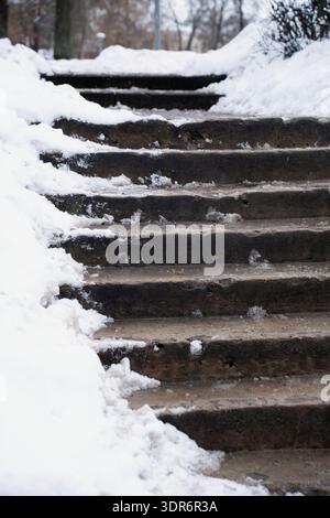 worn concrete outdoor staircase partially covered with fresh and melting snow, flanked by piled snowbanks in a quiet winter park, muted browns and whi Stock Photo