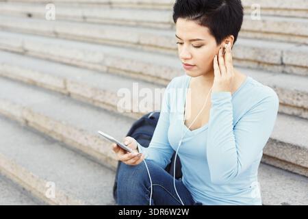 Woman Sits on Steps and Listens to Music on a Smartphone While Focusing on Her Screen Stock Photo
