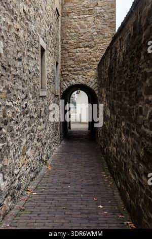 Narrow cobblestone alley framed by rough stone wall lead through arched passage toward light open courtyard. Medieval architecture texture shadow Stock Photo
