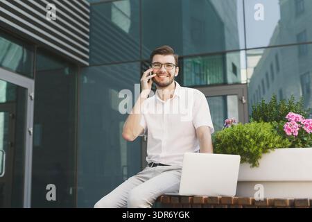 Man Sitting on a Table Outdoors Talking on the Phone and Working on a Laptop During the Day Stock Photo